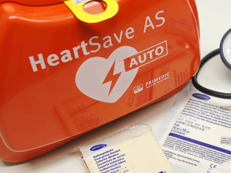 Das Bild zeigt einen Defibrillator in einer orangefarbenen Tasche neben einem Blutdruckmessgerät und mehreren verpackten Erste-Hilfe-Materialien.