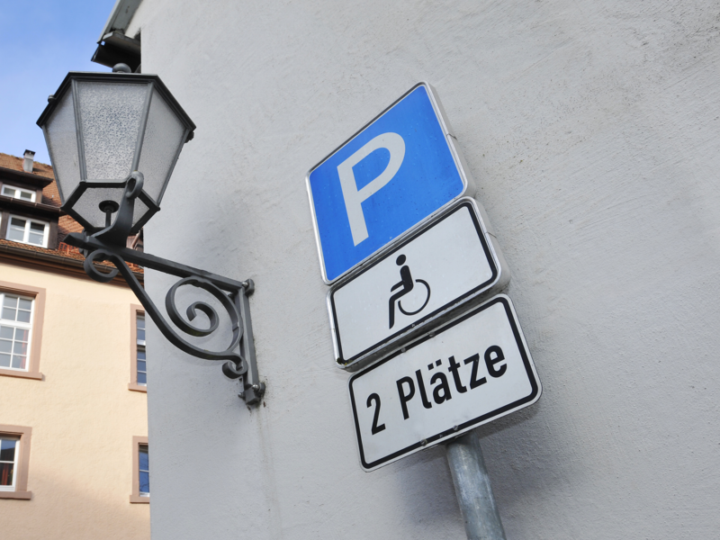 Ein Verkehrsschild für zwei Behindertenparkplätze ist neben einer antik aussehenden Straßenlaterne an einer Wand montiert, mit einem mehrstöckigen Gebäude im Hintergrund.