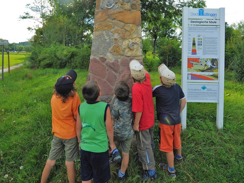 Eine Gruppe von Kindern steht vor einer geologischen Steinsäule in einem grünen Park und betrachtet diese aufmerksam.