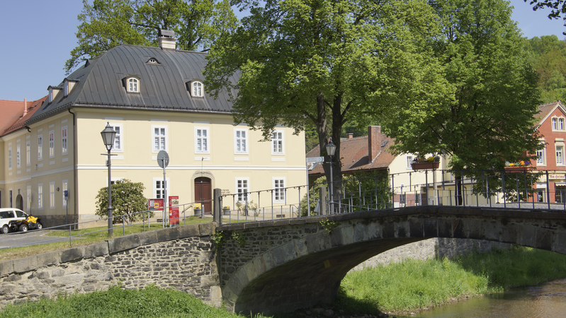 Das Kupferhaus in Großschönau - ein weiträumiges, gelbes Gebäude mit einem dunklen Dach - steht neben einer steinernen Bogenbrücke, flankiert von üppigen Bäumen.