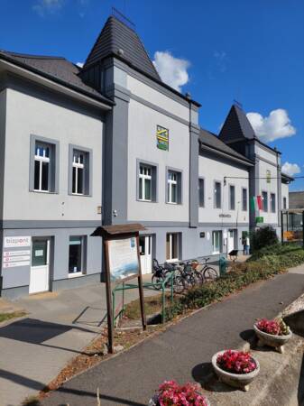 Das Bild zeigt ein großes, graues Gebäude in Mende mit spitzen Dächern und einem Wappen an der Fassade, umgeben von einem Bürgersteig mit Blumenbeeten und Fahrradständern.
