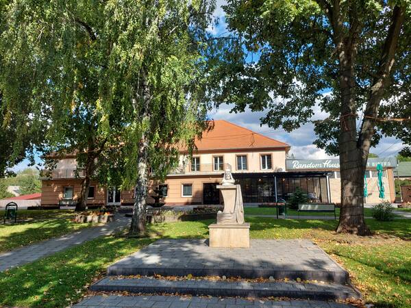 Ein kleines Gebäude in Mende mit roten Dachziegeln steht hinter einer Statue, umgeben von grünen Bäumen auf einem sonnigen Platz.