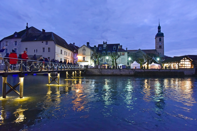 Dieses Bild zeigt eine Brücke in Ornans mit Weihnachtsbeleuchtung und voller Menschen, die über einen Fluss zu einer festlich erleuchteten Stadt mit historischen Gebäuden und einem Kirchturm führen.