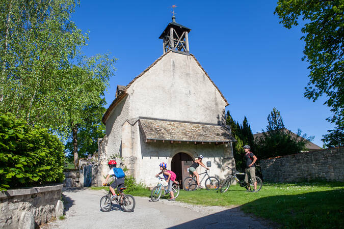 Eine Gruppe von Menschen mit Fahrrädern steht vor einer kleinen und alten Kirche unter dem blauen Himmel von Ornans.