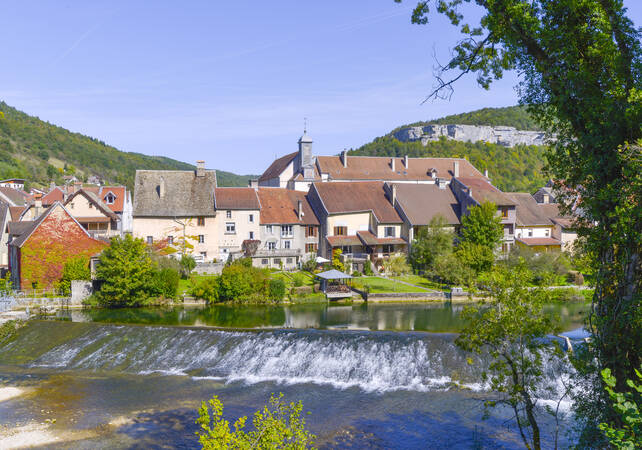 Unsere malerisches Partnerstadt Ornans mit traditionellen Häusern liegt am Ufer eines ruhigen Flusses, umgeben von grünen Hügeln.