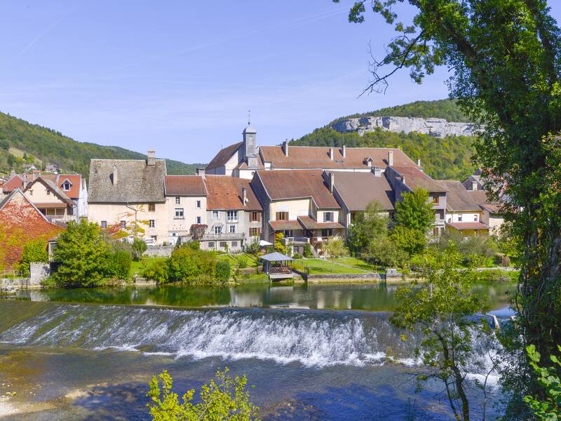 Unsere malerisches Partnerstadt Ornans mit traditionellen Häusern liegt am Ufer eines ruhigen Flusses, umgeben von grünen Hügeln.