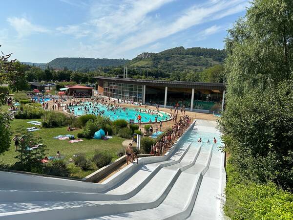 Das belebtes Freibad von Ornans mit mehreren Menschen im Wasser, umgeben von Grünflächen und einer großen Wasserrutsche im Vordergrund. Im Hintergrund befinden sich Hügeln und ein Wald.
