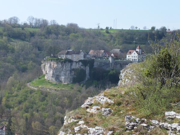 Eine kleine Siedlung in Ornans mit mehreren Gebäuden liegt malerisch auf einem steilen Felsvorsprung, umgeben von grünen Hügeln.