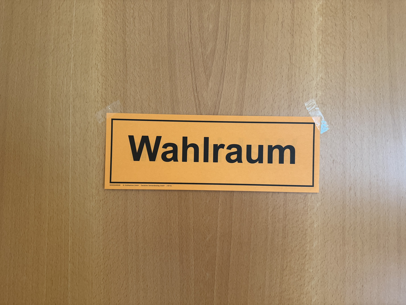 Auf diesem Bild ist eine Holztür zu sehen, an der ein orangefarbenes Schild mit der Aufschrift Wahlraum befestigt ist.