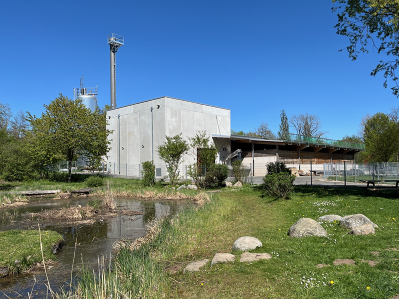 Ein modernes, graues Industriegebäude steht in einer grünen Landschaft mit Bäumen und einem kleinen Teich im Vordergrund.