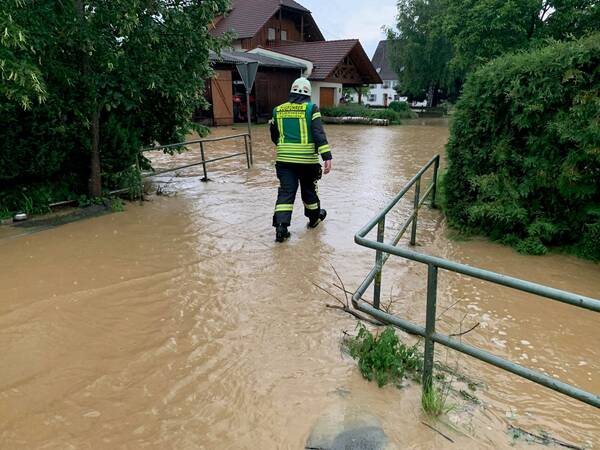 Ein Feuerwehrmann geht durch eine überflutete Straße vor einem Haus, umgeben von Bäumen und Sträuchern.
