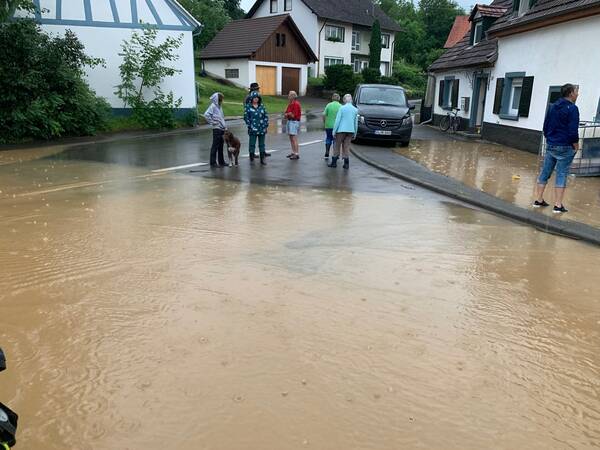 Mehrere Personen stehen auf einer überfluteten Straße in einem Wohngebiet mit nassen Autos und Häusern im Hintergrund.