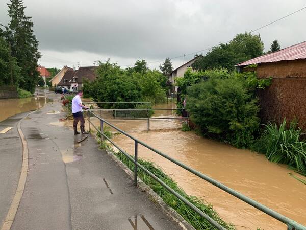 Eine überflutete Straße in einem Dorf mit braunem Wasser, das Häuser und Vegetation umgibt, während eine Person am Zaun steht.