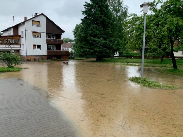 Eine Straße und ein kleines Wohngebiet sind vollständig von Hochwasser überschwemmt, wobei das Wasser bis an die Häuser und Bäume reicht.