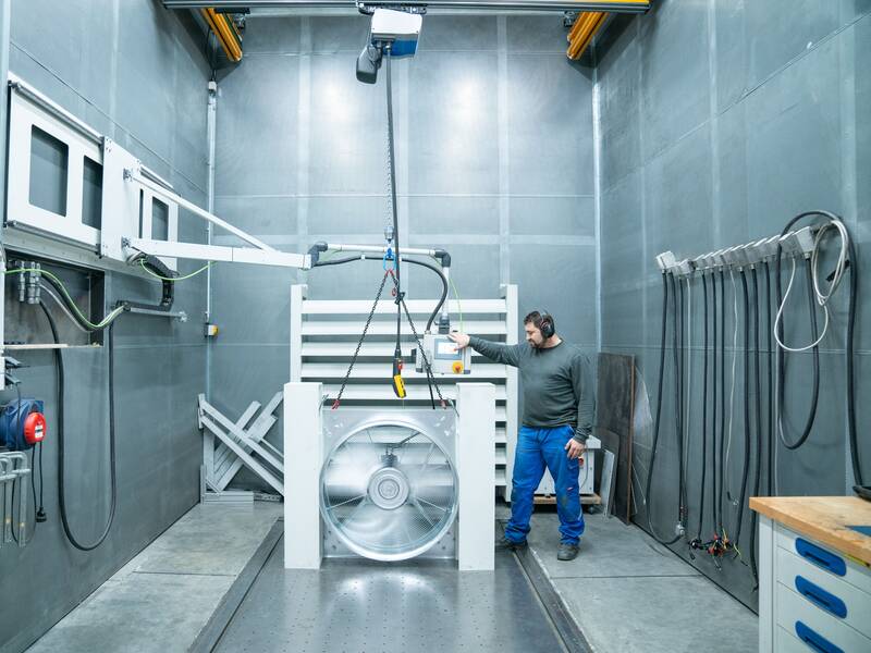 Ein Mann mit Gehörschutz steht in einem industriellen Testlabor und arbeitet mit einem großen Ventilator und technischen Geräten.