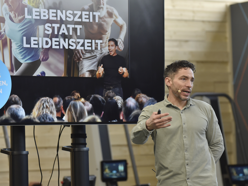 Ein Mann steht vor einem Publikum und einem Bildschirm während des Unternehmerfrühstücks 2026, der die Wörter "Lebenszeit statt Leidenszeit" und "Das Geheimnis eines langen, gesunden Lebens" zeigt.