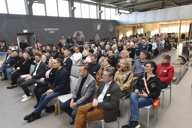 Eine Gruppe von Menschen sitzt in mehreren Reihen in einem modernen Konferenzraum und schaut aufmerksam nach vorne.