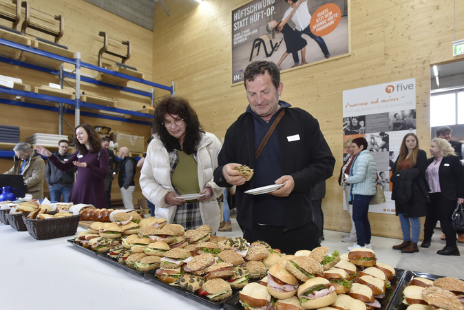 Menschen bedienen sich an einem Buffet mit verschiedenen belegten Brötchen in einem Raum mit Holzwänden und Regalen.