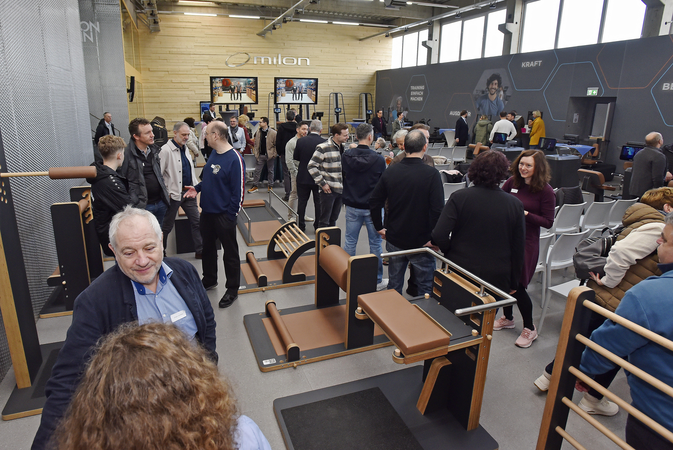 Eine Gruppe von Menschen versammelt sich in einem Ausstellungsraum voller Fitnessgeräte, während sie sich unterhalten und die Umgebung erkunden.