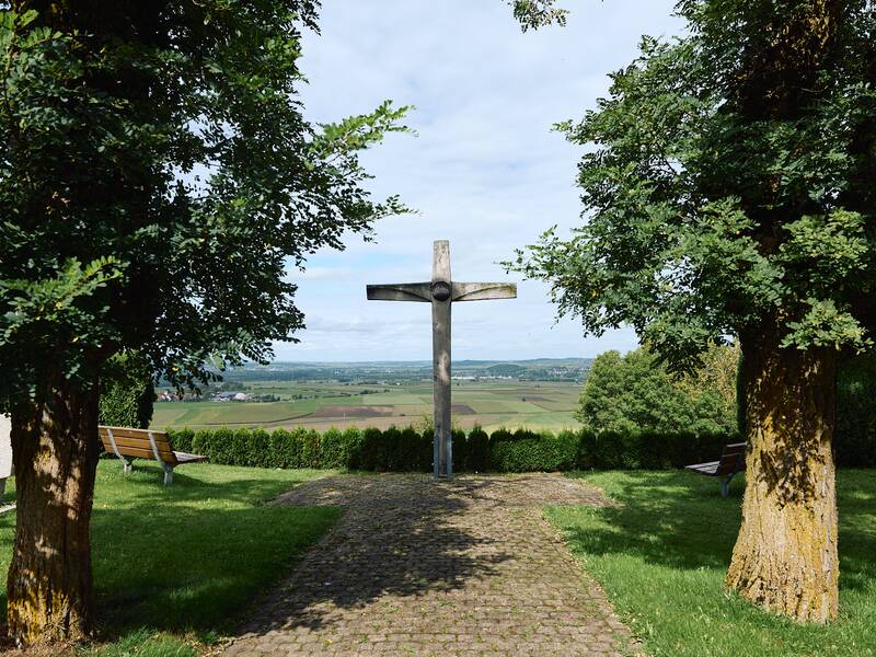 Ein großes Holzkreuz steht zwischen zwei Bäumen auf einem gepflasterten Weg mit Blick auf eine weitläufige Landschaft im Hintergrund.