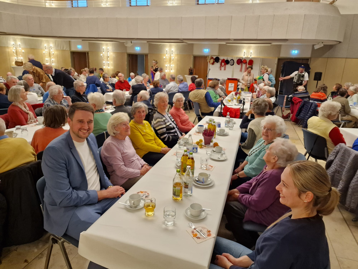 In einem hellen Saal sitzen viele ältere Menschen an langen, gedeckten Tischen, während im Hintergrund einige Personen in traditioneller Kleidung musizieren.