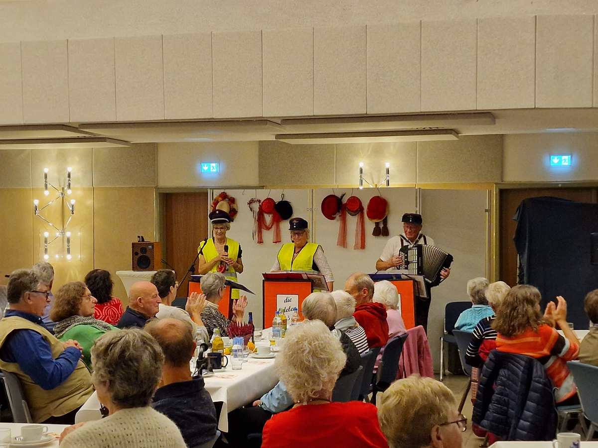 In der Stadthalle Hüfingen sitzen Menschen an gedeckten Tischen, während vorne drei Personen in gelben Westen und Hüten musizieren und sprechen.