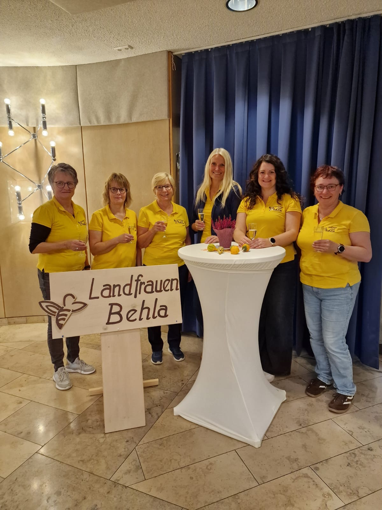 Sechs Landfrauen Behla stehen lächelnd um einen Stehtisch und tragen gelbe Hemden vor einem blauen Vorhang.