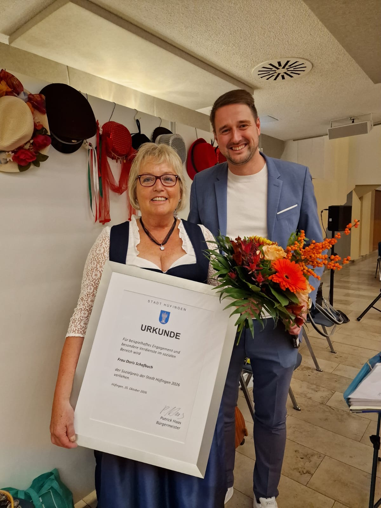 Doris Schafbuch hält eine Urkunde, während Bürgermeister Patrick Haas neben ihr steht und ihr einen Blumenstrauß überreicht; im Hintergrund sind Hüte zu sehen.