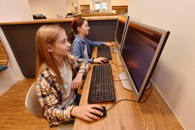 Zwei Kinder sitzen konzentriert an Computern in der Kinder- und Jugendbibliothek mit Holzboden und sind in Bildschirmarbeit vertieft.