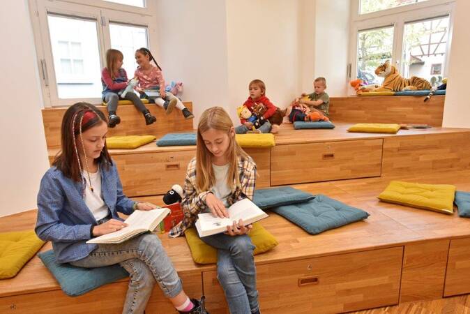 Auf einem mehrstufigen Holzpodest in der Kinder- und Jugendbibliothek sitzen mehrere Kinder auf bunten Kissen, während einige lesen und andere miteinander sprechen.