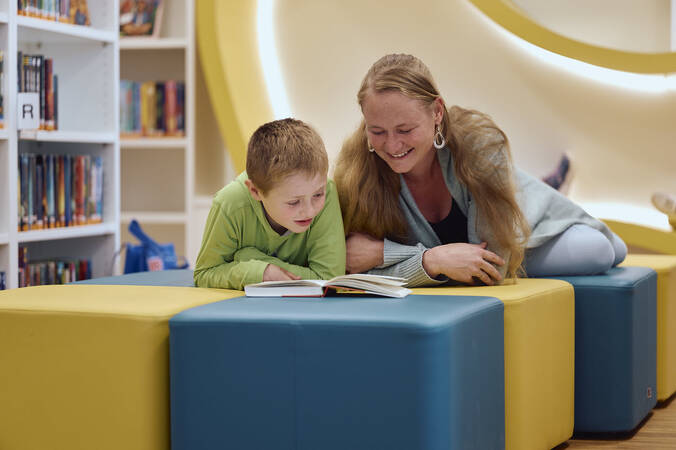 Ein Kind und eine Frau liegen auf farbigen Sitzwürfeln in der Kinder- und Jugendbibliothek und lesen gemeinsam ein Buch.
