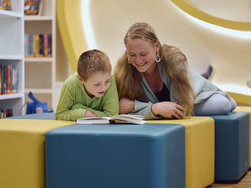 Ein Kind und eine Frau liegen auf farbigen Sitzwürfeln in der Kinder- und Jugendbibliothek und lesen gemeinsam ein Buch.