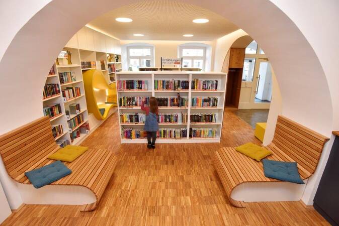 Ein Kind steht in der modernen und gut beleuchteten Kinder- und Jugendbibliothek umgeben von Bücherregalen und gemütlichen Sitzbänken aus Holz mit bunten Kissen.