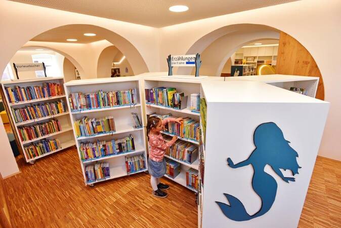 In der Kinder- und Jugendbibliothek mit Bücherregalen voller Kinderbücher auf denen eine Silhouette einer Meerjungfrau an der Seite eines Regals aufgebraucht ist, betrachtet ein Kind ein Buch.