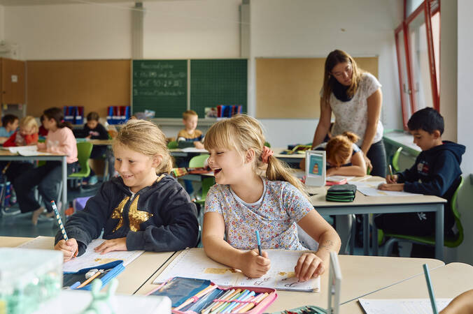 Eine Gruppe von Kindern sitzt an Schultischen im Klassenzimmer der Schellenberger Schule und arbeitet, während eine Lehrerin einem Schüler hilft.