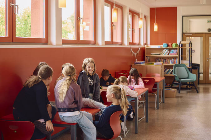 Im Flur der Schellenberger Schule sitzen mehrere Kinder an Tischen und unterhalten sich fröhlich, während andere schreiben, umgeben von Regalen mit Büchern.