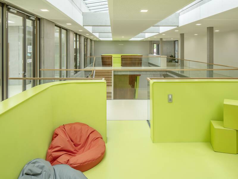 Ein moderner Innenraum der Lucian-Reich-Schule mit hellgrünen Wänden und Boden, Sitzsäcken und gepolsterten Würfeln, sowie einem Glasgeländer und großen Fenstern auf der linken Seite.