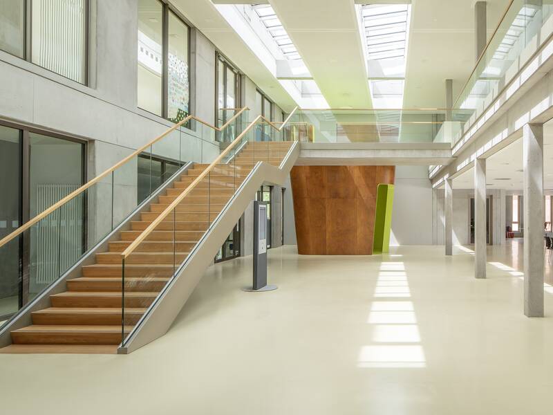 Der helle, moderne Innenraum der Lucian-Reich-Schule mit einer breiten Treppe aus Holz, einem Oberlicht und großen Fenstern, die den Raum mit Licht durchfluten.