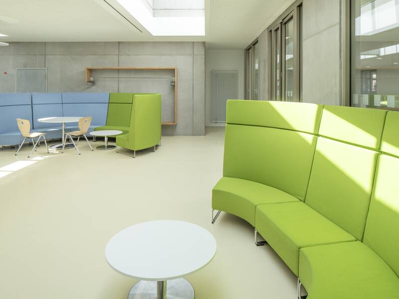 Ein moderner, heller Raum der Lucian-Reich-Schule mit geschwungenen Sofas in Grün und Blau, Tischgruppen und großen Fenstern, die viel Licht hineinlassen.