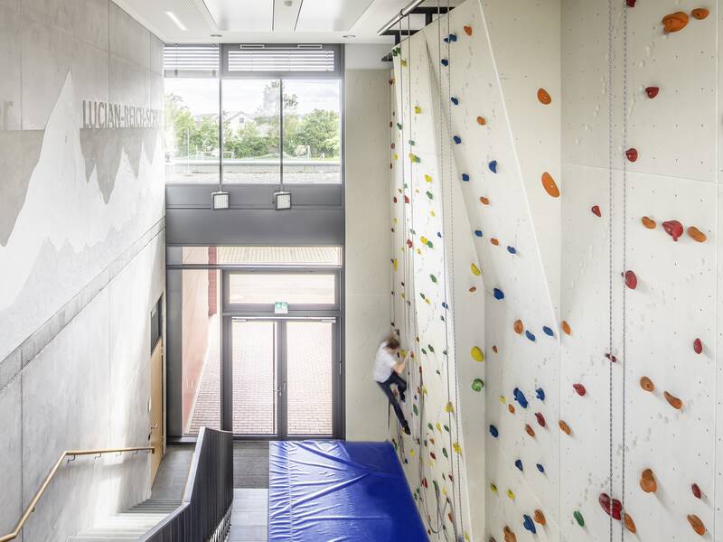 Eine Person klettert an einer bunten Indoor-Kletterwand, die sich in dem modernen, lichtdurchfluteten Flur der Lucian-Reich-Schule mit Treppe und blauen Sicherheitsmatten befindet.