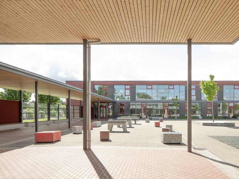 Der Schulhof ist mit gepflasterten Wegen, bunten Sitzblöcken und überdachten Bereichen gestaltet und liegt vor einem modernen Gebäude mit großen Fenstern.