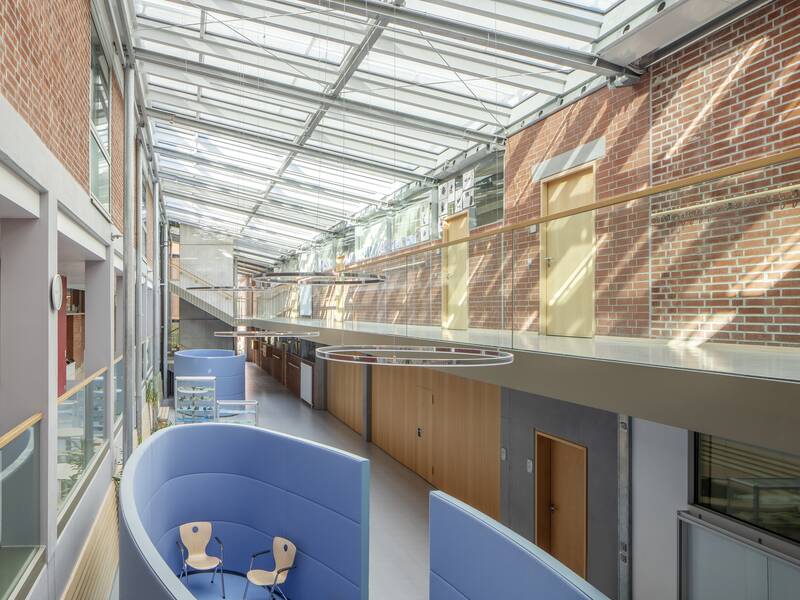 Ein heller, moderner Innenraum mit einer Glasdecke, roten Ziegelwänden, einem offenen Treppenhaus und blauen Sitzgelegenheiten.