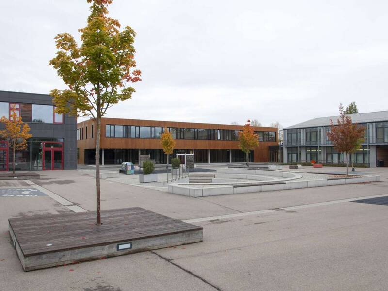 Der moderne Schulhof der Lucian-Reich-Schule mit grauen und roten Schulgebäuden, einigen Bäumen und einem zentralen, kreisförmigen Sitzbereich aus Beton.