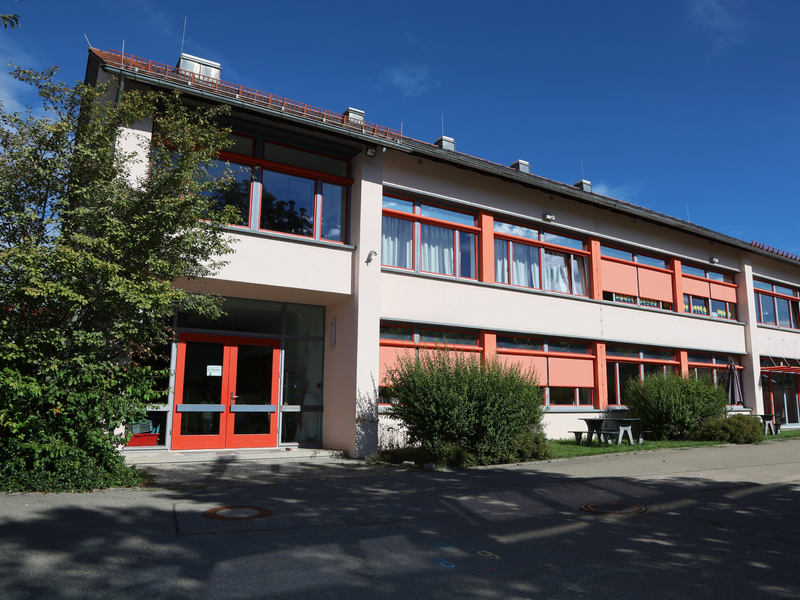 Das Bild zeigt die Schellenberger Schule in Hausen vor Wald mit roten Fenstern und Türen sowie einem flachen Dach, umgeben von Büschen und einem Baum, bei blauem Himmel.