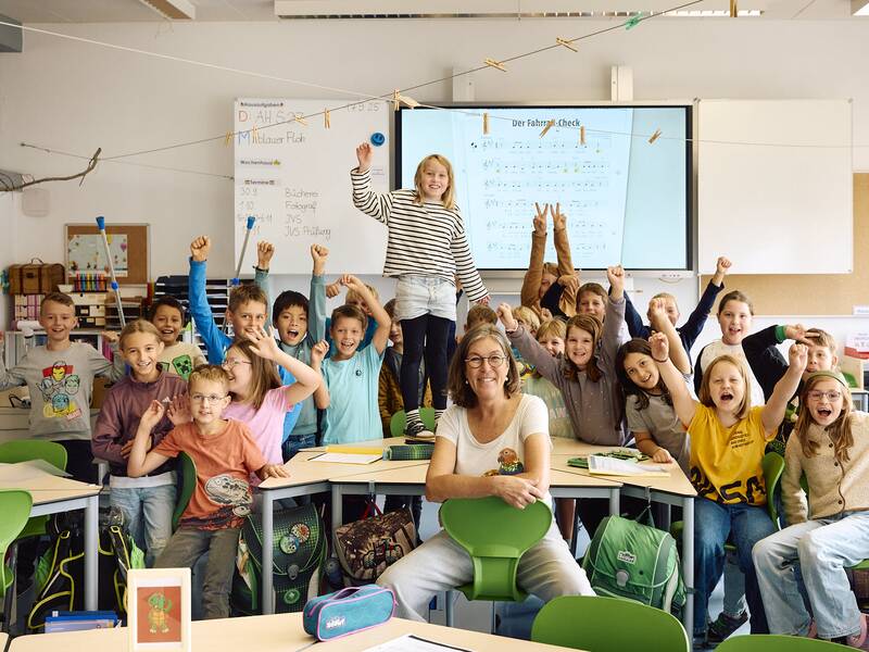 Eine Gruppe von fröhlichen Schulkindern und einer Lehrerin sitzt im Klassenzimmer der Schellenberger Schule mit der interaktiven Tafel im Hintergrund. Viele Kinder recken jubelnd die Fäuste in die Luft.