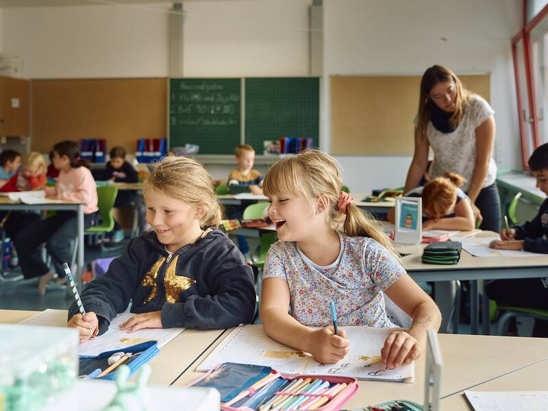 Eine Gruppe von Kindern sitzt an Schultischen im Klassenzimmer der Schellenberger Schule und arbeitet, während eine Lehrerin einem Schüler hilft.