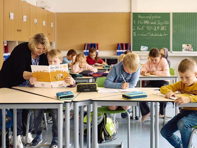 In einem Klassenzimmer der Schellenberger Schule arbeiten Kinder konzentriert an ihren Aufgaben, während eine Lehrerin einem Schüler hilft.