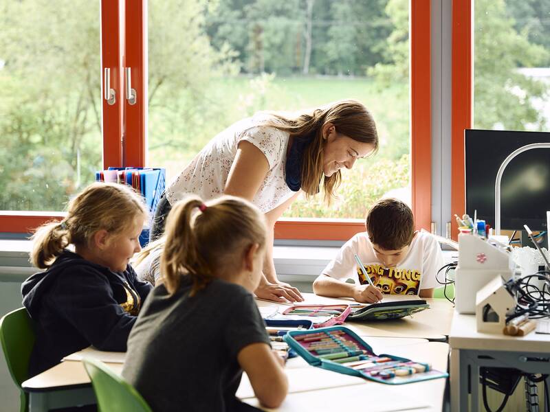 Eine Lehrerin beugt sich lächelnd zu einem Schüler, während zwei weitere Kinder in einem hellen Klassenzimmer an ihren Tischen sitzen und arbeiten.