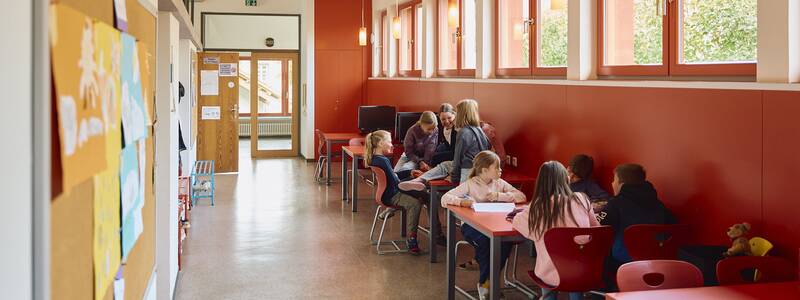 Im Flur der Schellenberger Schule sitzen mehrere Kinder an Tischen und unterhalten sich fröhlich, während andere schreiben, umgeben von Regalen mit Büchern.