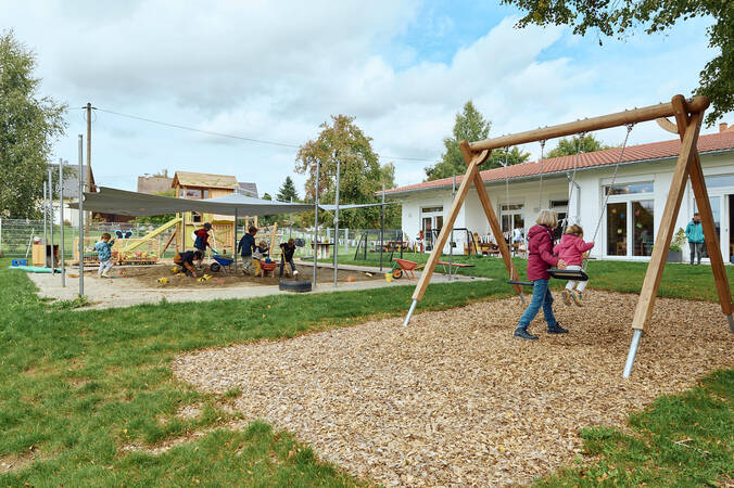 Im Außenbereich des Kindergartens schaukeln zwei Kinder, während andere im Sandkasten spielen.